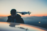 A drone is in focus, hovering in the air with blurred background elements. A person wearing a beanie and jacket is slightly out of focus, standing behind the drone. The setting appears to be during sunset or sunrise, with a gradient sky ranging from orange to blue.