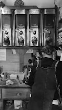 A person wearing an apron stands in front of a set of four large coffee dispensers labeled with different coffee types including Colombia and Guatemala. The area appears to be a coffee preparation station with various tools and containers on the counter.