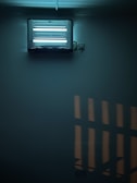 A fluorescent insect trap mounted on a wall. The device is emitting a bluish light, creating a soft glow in the surrounding area. The light casts geometric shadows on the wall, contributing to a slightly moody atmosphere.