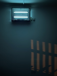 A fluorescent insect trap mounted on a wall. The device is emitting a bluish light, creating a soft glow in the surrounding area. The light casts geometric shadows on the wall, contributing to a slightly moody atmosphere.