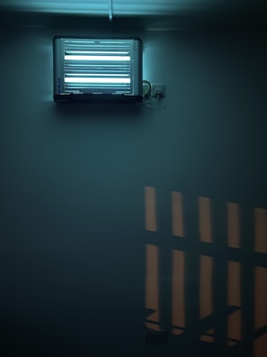 A fluorescent insect trap mounted on a wall. The device is emitting a bluish light, creating a soft glow in the surrounding area. The light casts geometric shadows on the wall, contributing to a slightly moody atmosphere.