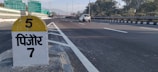 A highway scene with a milestone indicating '5' and the name of a location in Hindi script. The road is wide and mostly empty, with a white car traveling in the distance. Signboards with directions are visible overhead, and there are trees lining the sides. Mountainous terrain is faintly visible in the background.