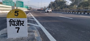 A highway scene with a milestone indicating '5' and the name of a location in Hindi script. The road is wide and mostly empty, with a white car traveling in the distance. Signboards with directions are visible overhead, and there are trees lining the sides. Mountainous terrain is faintly visible in the background.