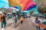 A vibrant outdoor scene with a colorful canopy hovering above a group of people gathered in an open plaza. Various individuals are engaged in different activities; some are standing, talking, or using their phones. The environment appears lively with children and adults interacting in a casual setting. Trees with autumn leaves frame the area, and urban buildings are visible in the background.