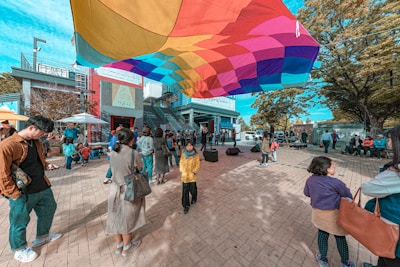 A vibrant outdoor scene with a colorful canopy hovering above a group of people gathered in an open plaza. Various individuals are engaged in different activities; some are standing, talking, or using their phones. The environment appears lively with children and adults interacting in a casual setting. Trees with autumn leaves frame the area, and urban buildings are visible in the background.