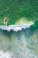 A lone surfer paddles through clear, green ocean waters towards an approaching white wave. The aerial perspective highlights the texture of the water, revealing intricate patterns of foam and contrasting colors.