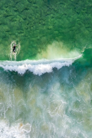 A lone surfer paddles through clear, green ocean waters towards an approaching white wave. The aerial perspective highlights the texture of the water, revealing intricate patterns of foam and contrasting colors.