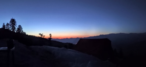 A serene mountain landscape at dusk with silhouetted trees and a building against a gradient sky transitioning from deep blue to a warm orange near the horizon, suggesting the setting or rising of the sun.