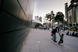 A modern urban scene depicting a large, curved architectural structure with a smooth exterior. People are casually walking and interacting on a spacious, paved area. Tall buildings and buses are visible in the background with trees lining the pathway. The sky appears to be during dusk or dawn, offering a serene urban ambiance.