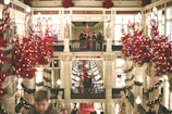 An ornate shopping gallery adorned with festive red decorations and poinsettias. Statues and columns are visible, and people are walking around, creating a lively holiday atmosphere.