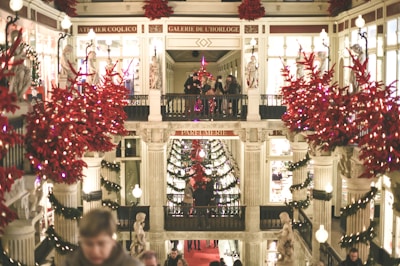 An ornate shopping gallery adorned with festive red decorations and poinsettias. Statues and columns are visible, and people are walking around, creating a lively holiday atmosphere.