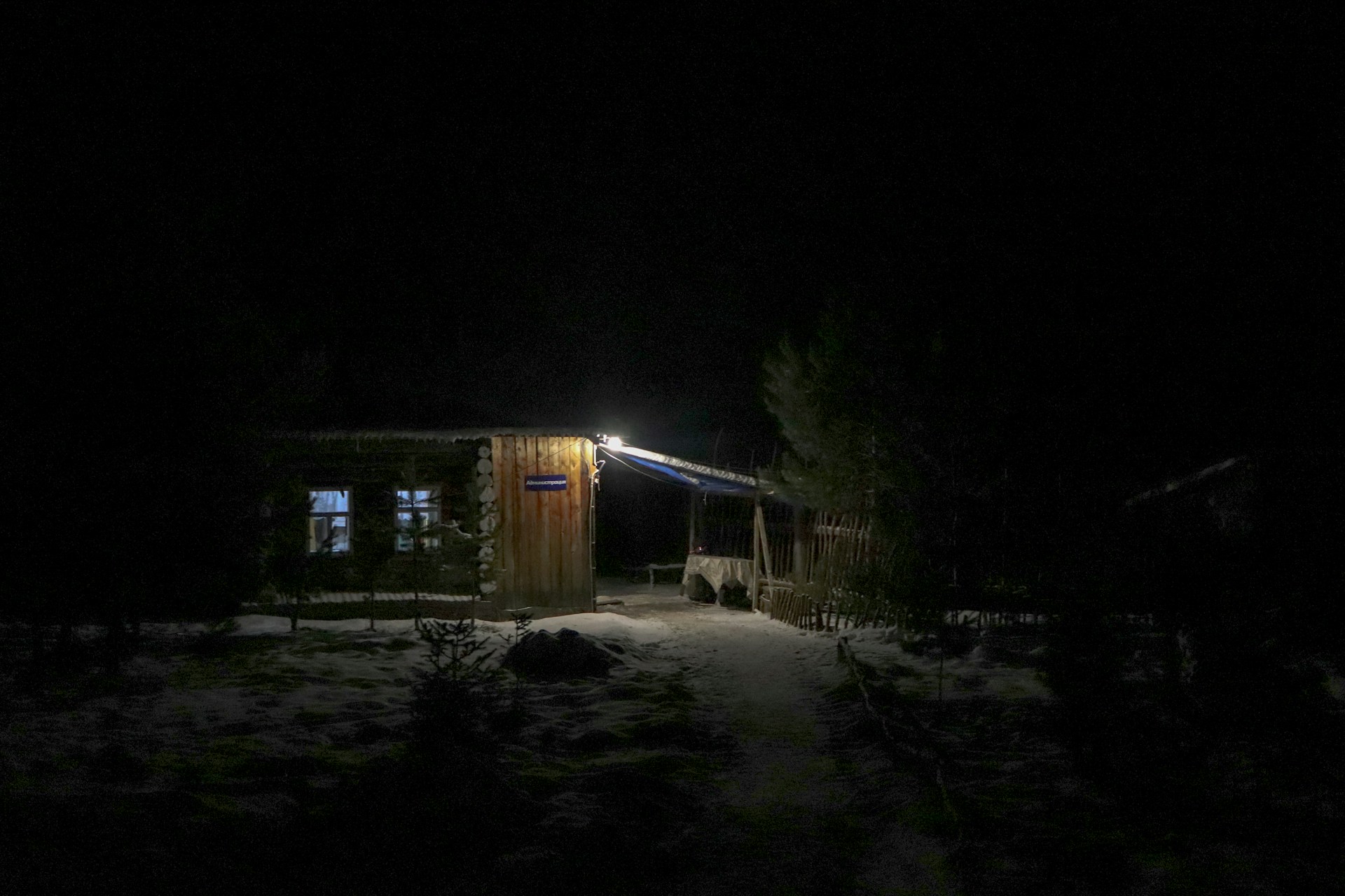An abandoned, dimly lit cabin window with faint silhouettes visible inside, framed by twisted branches.