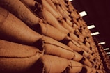 A large stack of burlap sacks arranged neatly in a warehouse setting, with a warm, sepia-toned lighting that gives an industrial and rustic appearance.