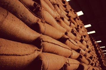 A large stack of burlap sacks arranged neatly in a warehouse setting, with a warm, sepia-toned lighting that gives an industrial and rustic appearance.