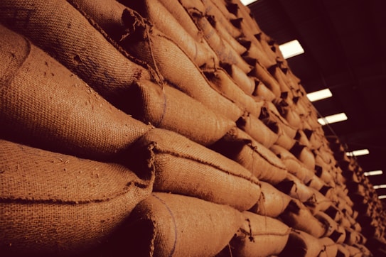 A large stack of burlap sacks arranged neatly in a warehouse setting, with a warm, sepia-toned lighting that gives an industrial and rustic appearance.