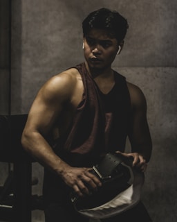 A muscular individual is in a dimly lit environment, likely a gym. They are wearing a sleeveless shirt and holding a weightlifting belt. Their expression appears focused and intense.
