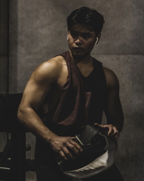 A muscular individual is in a dimly lit environment, likely a gym. They are wearing a sleeveless shirt and holding a weightlifting belt. Their expression appears focused and intense.