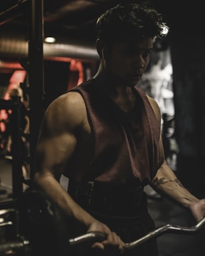 A person is intensely focused while lifting weights in a dimly lit gym. The muscular individual is wearing a sleeveless shirt and earphones, indicating concentration on their workout. The background is blurred, showcasing gym equipment and a soft glow of red and orange lights.