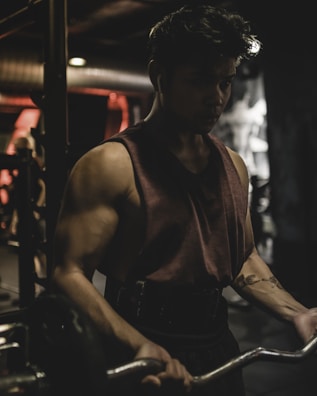 A person is intensely focused while lifting weights in a dimly lit gym. The muscular individual is wearing a sleeveless shirt and earphones, indicating concentration on their workout. The background is blurred, showcasing gym equipment and a soft glow of red and orange lights.