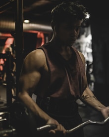 A person is intensely focused while lifting weights in a dimly lit gym. The muscular individual is wearing a sleeveless shirt and earphones, indicating concentration on their workout. The background is blurred, showcasing gym equipment and a soft glow of red and orange lights.