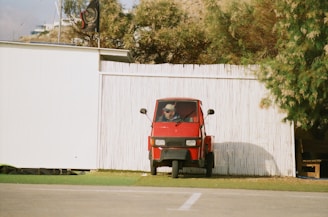 A small red three-wheeled vehicle is parked on grass beside a white wooden fence. A person is seated inside the vehicle, wearing sunglasses and a hat. The background features green leafy trees and a building with a flat roof and flags.
