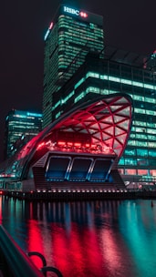 A modern cityscape at night featuring illuminated skyscrapers with bright logos, a futuristic building with a canopy reflecting vibrant neon lights in the water.
