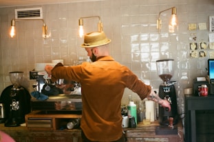 A barista wearing a straw hat and brown shirt prepares a coffee drink at an espresso machine in a cafe. The setting has a rustic vibe with a tiled wall, exposed hanging light bulbs, and various coffee-making tools and ingredients on the counter.