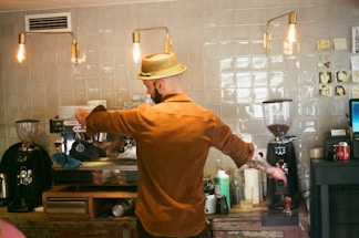 A barista wearing a straw hat and brown shirt prepares a coffee drink at an espresso machine in a cafe. The setting has a rustic vibe with a tiled wall, exposed hanging light bulbs, and various coffee-making tools and ingredients on the counter.