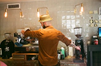 A barista wearing a straw hat and brown shirt prepares a coffee drink at an espresso machine in a cafe. The setting has a rustic vibe with a tiled wall, exposed hanging light bulbs, and various coffee-making tools and ingredients on the counter.
