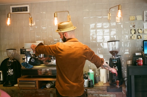 A barista wearing a straw hat and brown shirt prepares a coffee drink at an espresso machine in a cafe. The setting has a rustic vibe with a tiled wall, exposed hanging light bulbs, and various coffee-making tools and ingredients on the counter.