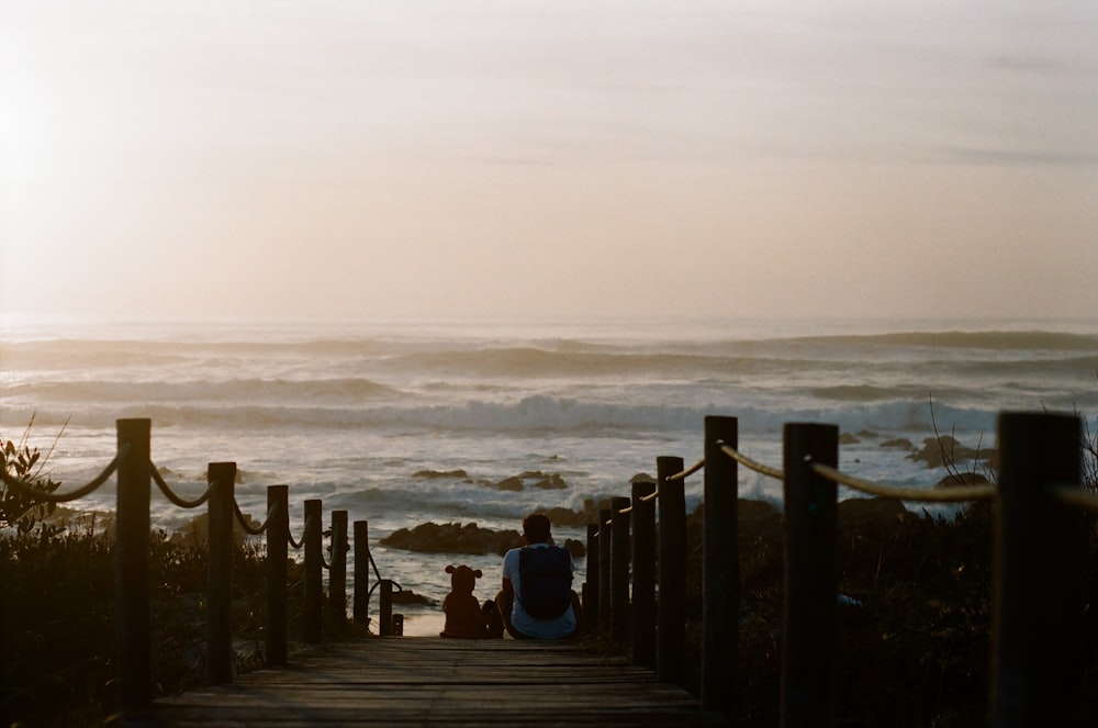 man and sitting on wooden dock beside dog facing ocean