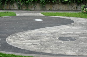 A large circular yin-yang symbol made of stones is embedded into a paved surface. The contrasting black and white areas are distinct, with a small circle of each color within the opposite side. The surrounding area includes green grass and a wall covered with vines.