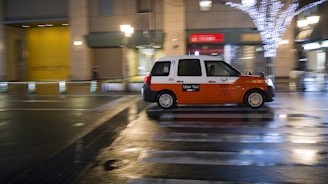 A taxi with an orange and white color scheme is moving quickly down a city street at night. The street is wet, reflecting lights from nearby buildings and street lamps, including a tree adorned with white string lights. The scene suggests a bustling urban environment.