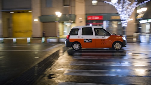 A taxi with an orange and white color scheme is moving quickly down a city street at night. The street is wet, reflecting lights from nearby buildings and street lamps, including a tree adorned with white string lights. The scene suggests a bustling urban environment.