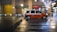 A taxi with an orange and white color scheme is moving quickly down a city street at night. The street is wet, reflecting lights from nearby buildings and street lamps, including a tree adorned with white string lights. The scene suggests a bustling urban environment.