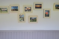 A wall decorated with eight framed photographs displayed in two horizontal rows. The photographs feature various subjects, including landscapes, mushrooms, wheels, and a mushroom cap. The frames are slightly varied in color but share a similar vintage style. Underneath the frames, there is a light gray wooden paneling.