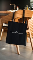A black tote bag with a minimalist design hangs on the back of a wooden chair in a modern kitchen setting. The kitchen features a wooden dining table, kitchen appliances in the background, and a warm, ambient lighting.