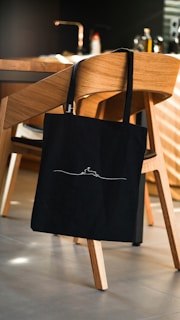 A black tote bag with a minimalist design hangs on the back of a wooden chair in a modern kitchen setting. The kitchen features a wooden dining table, kitchen appliances in the background, and a warm, ambient lighting.