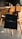 A black tote bag with a minimalist design hangs on the back of a wooden chair in a modern kitchen setting. The kitchen features a wooden dining table, kitchen appliances in the background, and a warm, ambient lighting.