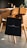 A black tote bag with a minimalist design hangs on the back of a wooden chair in a modern kitchen setting. The kitchen features a wooden dining table, kitchen appliances in the background, and a warm, ambient lighting.