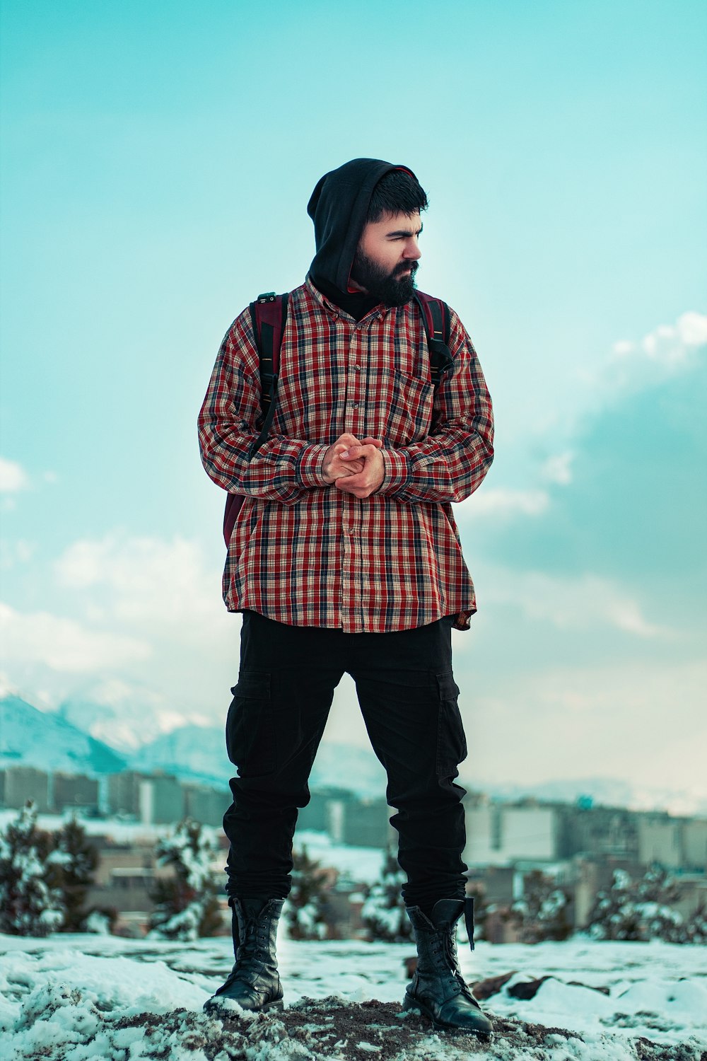 man wearing red, brown, and black plaid collared button-up long-sleeved shirt standing while glancing his left side under blue and white sky