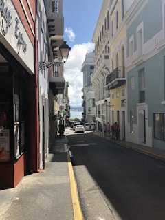 A narrow street lined with colonial-style buildings featuring colorful facades in shades of red, yellow, and blue. Ornate street lamps hang from the buildings. The street leads downhill and is busy with pedestrians, showcasing a lively urban environment. In the distance, the sea is visible under a clear blue sky.