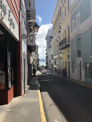 A narrow street lined with colonial-style buildings featuring colorful facades in shades of red, yellow, and blue. Ornate street lamps hang from the buildings. The street leads downhill and is busy with pedestrians, showcasing a lively urban environment. In the distance, the sea is visible under a clear blue sky.
