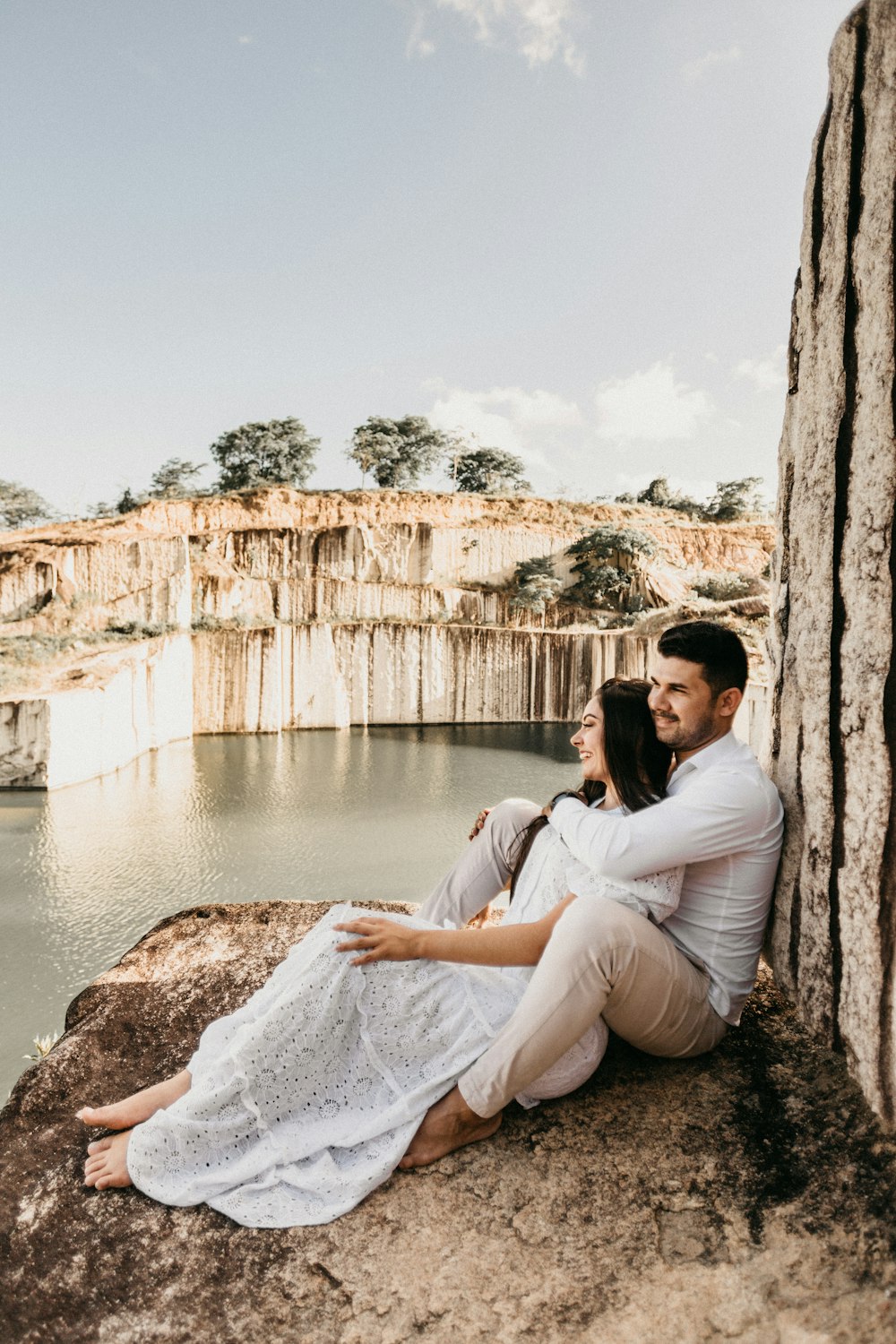 smiling man and woman sitting on rock near body of water during daytime