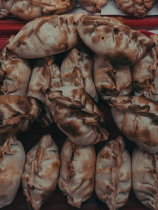 A close-up view of a pile of baked goods, likely empanadas, featuring a golden-brown crust with slightly darker patches. Each pastry is folded and crimped at the edges, showcasing an artisanal and homemade appearance.