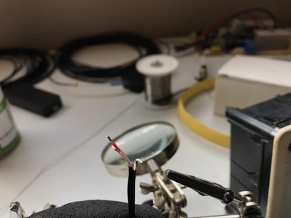 A workspace with a focus on a soldering setup where a helping hand tool is holding a wire. The wire has exposed ends and is positioned in front of a magnifying glass. In the background, various spools, cables, and electronic components are spread on the table.