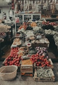 A bustling market stall richly filled with a diverse array of fresh fruits and vegetables. Crates of ripe red tomatoes are prominently displayed at the front alongside baskets of carrots, eggplants, bananas, and beans. The background is lined with shelves stocked with various packaged goods, and refrigerated cases are visible, adding to the vibrant array of food items.