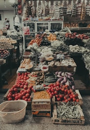 A bustling market stall richly filled with a diverse array of fresh fruits and vegetables. Crates of ripe red tomatoes are prominently displayed at the front alongside baskets of carrots, eggplants, bananas, and beans. The background is lined with shelves stocked with various packaged goods, and refrigerated cases are visible, adding to the vibrant array of food items.