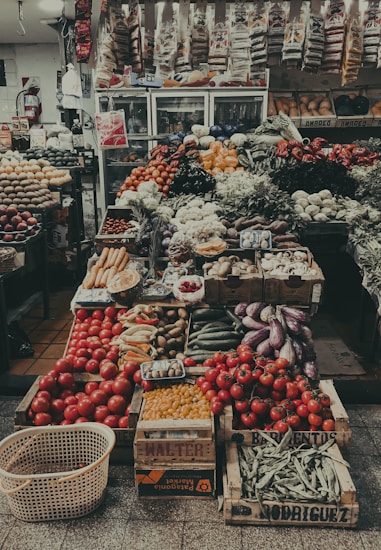 A bustling market stall richly filled with a diverse array of fresh fruits and vegetables. Crates of ripe red tomatoes are prominently displayed at the front alongside baskets of carrots, eggplants, bananas, and beans. The background is lined with shelves stocked with various packaged goods, and refrigerated cases are visible, adding to the vibrant array of food items.