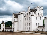 A historic white castle with multiple towers and crenellated parapets stands under a cloudy sky. The building features a Scottish flag on a pole, dark roof tiles, and small windows. Black cannons are lined up on the gravel courtyard alongside the building. Two people are visible near the entrance of a smaller tower with a clock.